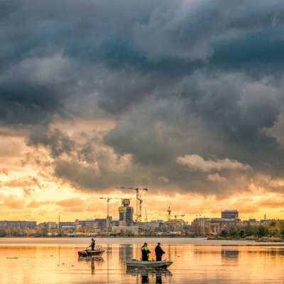 A body of water with several small boats or fishing vessels on the surface. In the background, there is a cityscape with a mix of modern high-rise buildings and older structures, including a smokestack emitting white smoke