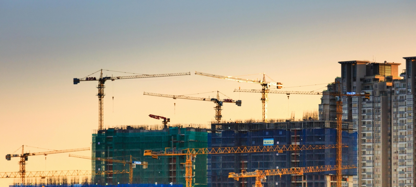 A cityscape of high-rise buildings under construction, wrapped in green and blue mesh, with numerous tower cranes operating at sunset.