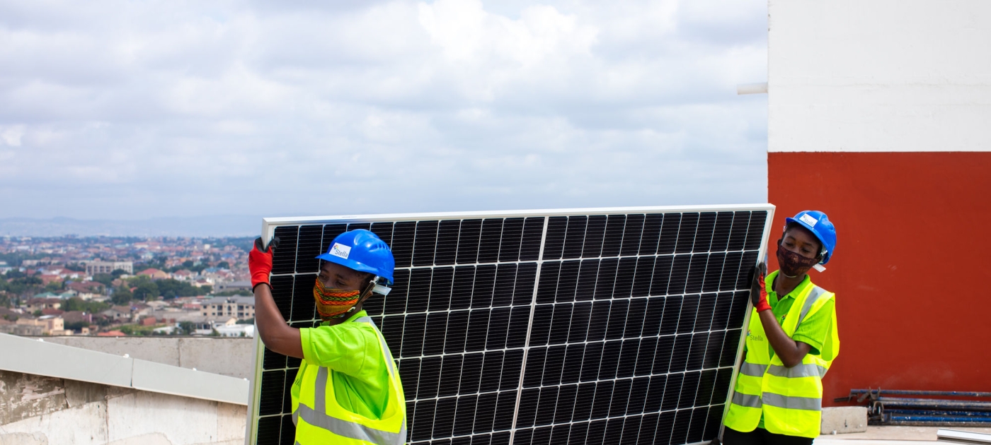 Two women wering workimng vests transport a solar panel in a rooftop 