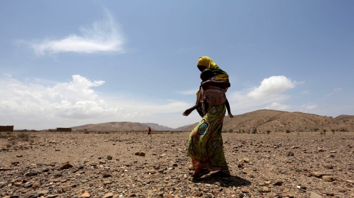 A woman walks across a dusty field with a baby.