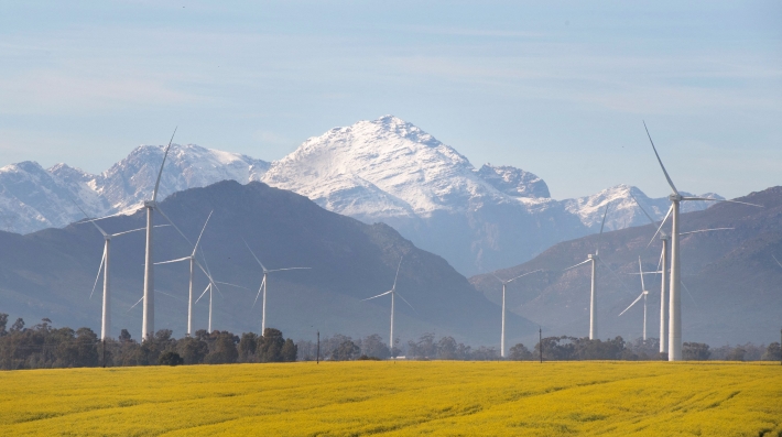 Several wind turbines overlook a field with snow-capped mountains in the background 