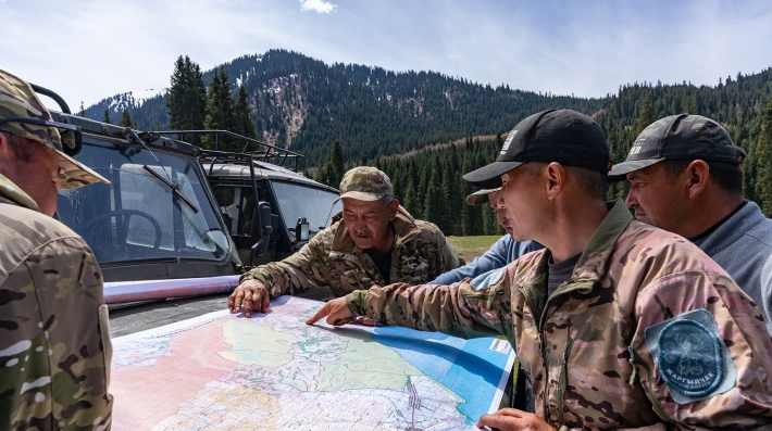 Cinco hombres estudian un mapa, con grandes montañas rodeándoles