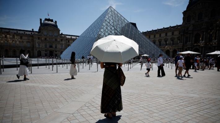 Una persona de pie bajo una sombrilla, frente a la pirámide de cristal del museo del Louvre en París.