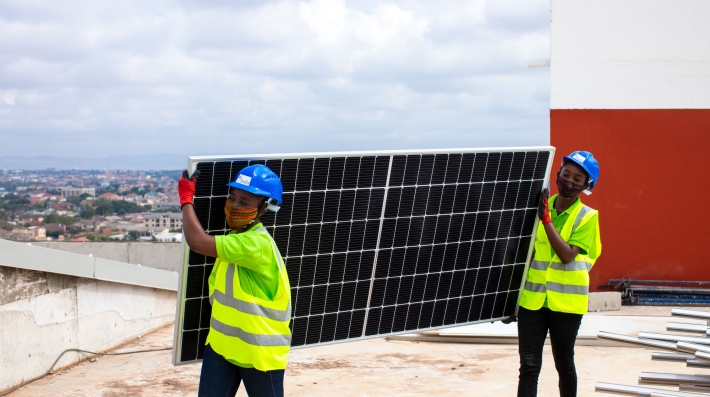 Two women wering workimng vests transport a solar panel in a rooftop 